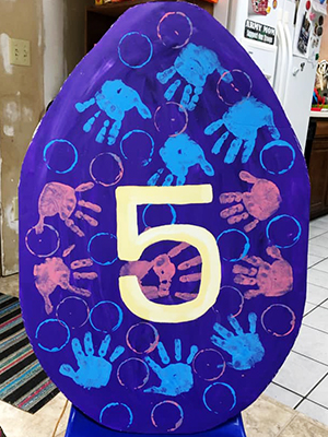 A sign in the shape of a large dark blue egg, decorated with lighter blue and pink handprints and the number 5.