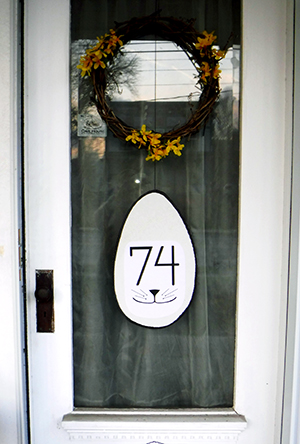Front door with a spring wreath above a large white egg with bunny whiskers and the number 74.