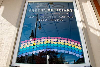 Store window of Sheehy Opticians with a rainbow where each row of color is made from colored cutouts in the shape of eyeglases.