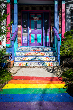 Entrance to a colorful Victorian house, with a rainbow painted on the walkway and in mosaic on the stair risers.