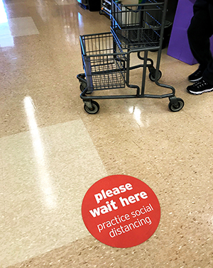 Round sign on the floor of a grocery store asking customers in line to 'please wait here/practice social distancing'