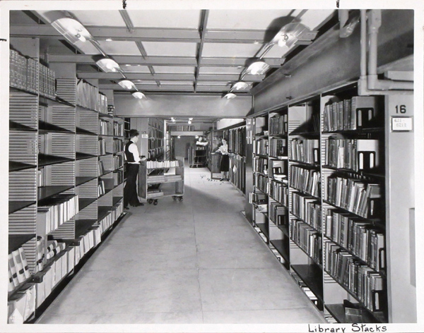 Library stacks in the 1940s, with shelves of books and a man with a book cart in the distance.