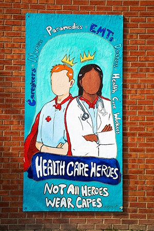 Sign on brick wall with two healthcare workers: Health Care Heroes - Not all heroes wear capes.