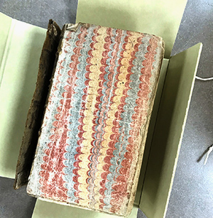 archival box open to show an old book with the cover missing; the page visible is marbled in red, blue and yellow